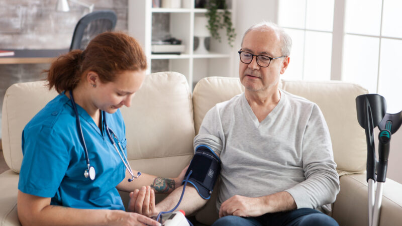 Female doctor reading blood pressure of old man in nursing home using digital device. Nurse and patient sitting on couch.